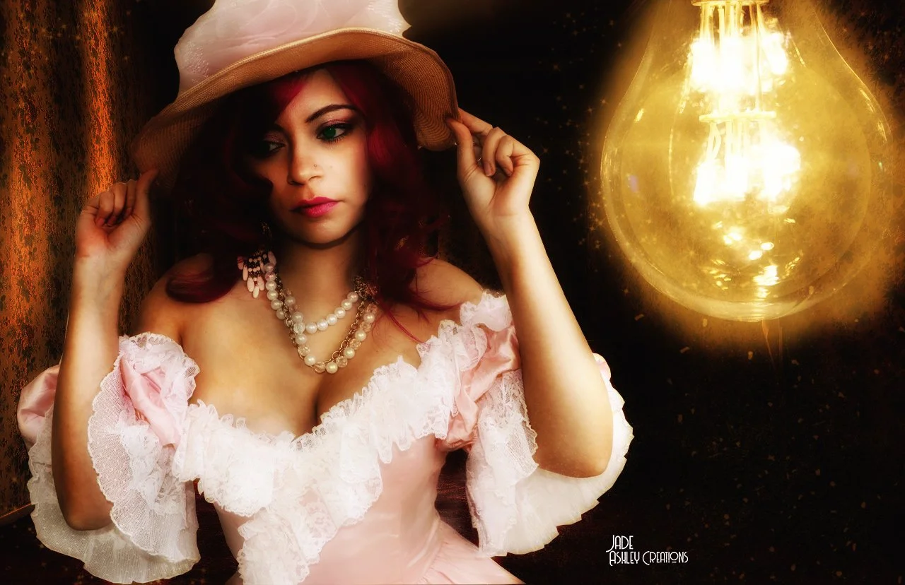 A woman with red hair wearing a pink, off-shoulder dress with lace trim, a pearl necklace, and a wide-brimmed hat, touching her hat with one hand, standing near a glowing vintage light bulb against a dark backdrop.