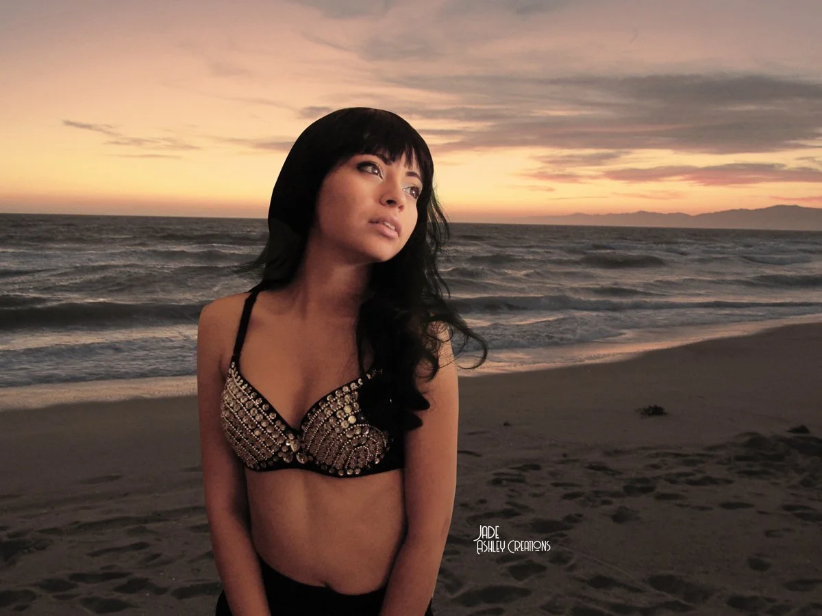 A woman with black hair and bangs wearing a studded black bra, standing on a beach during sunset with the ocean in the background.