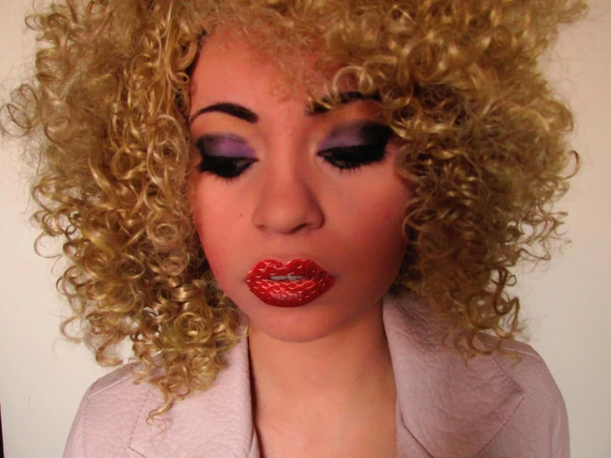 Close-up of a woman with blonde, curly hair, bold eye makeup, and shiny red lipstick, wearing a light pink jacket.