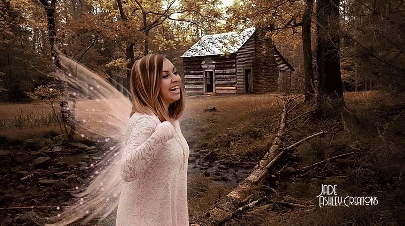 A woman smiling and wearing fairy wings stands in a forest clearing near a small log cabin, with autumn foliage in the background.