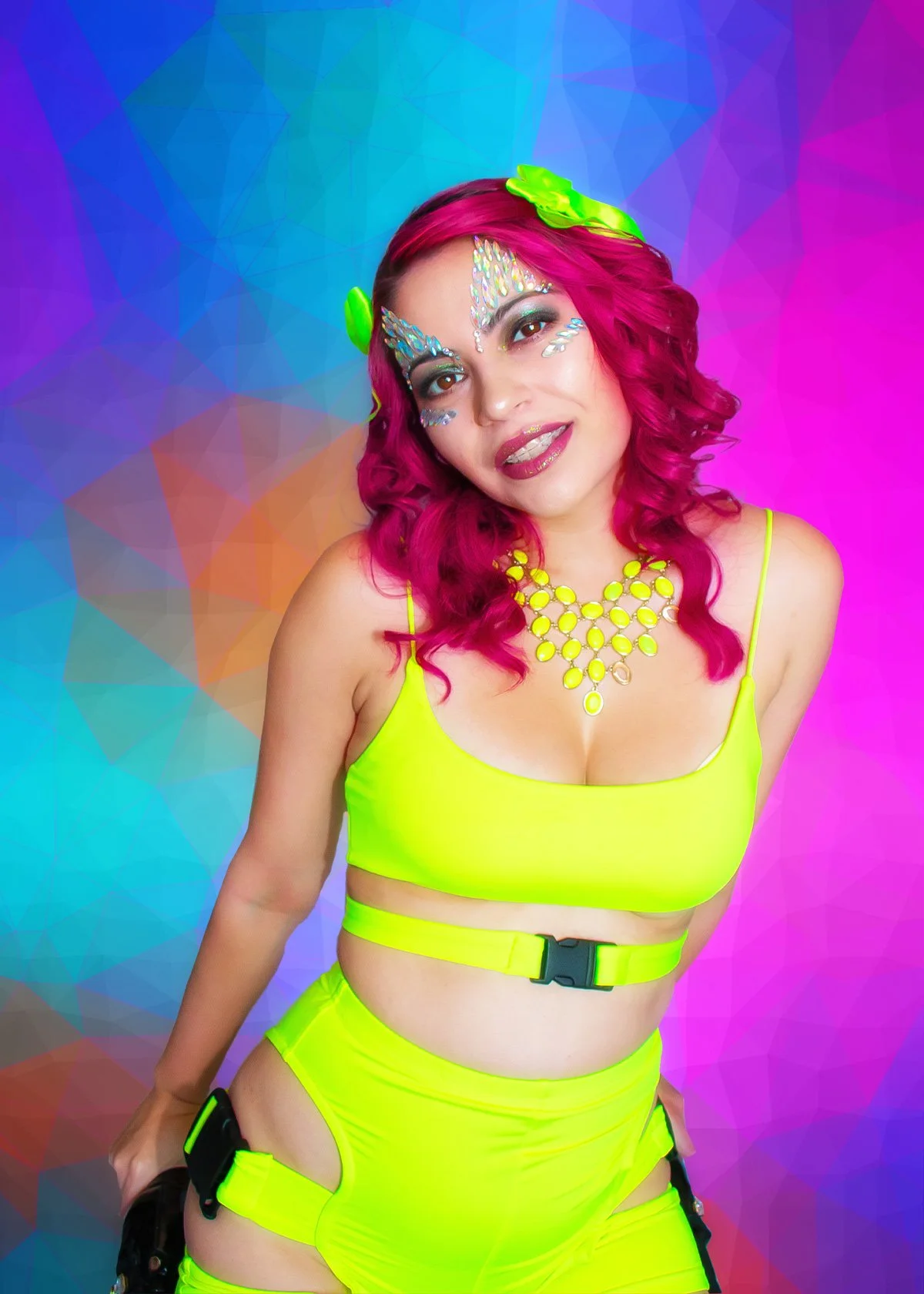 A woman with bright pink hair styled in loose waves, wearing a neon yellow crop top and matching shorts with cutouts and straps. She has colorful makeup with holographic and rhinestone embellishments on her face, and is accessorized with a statement 