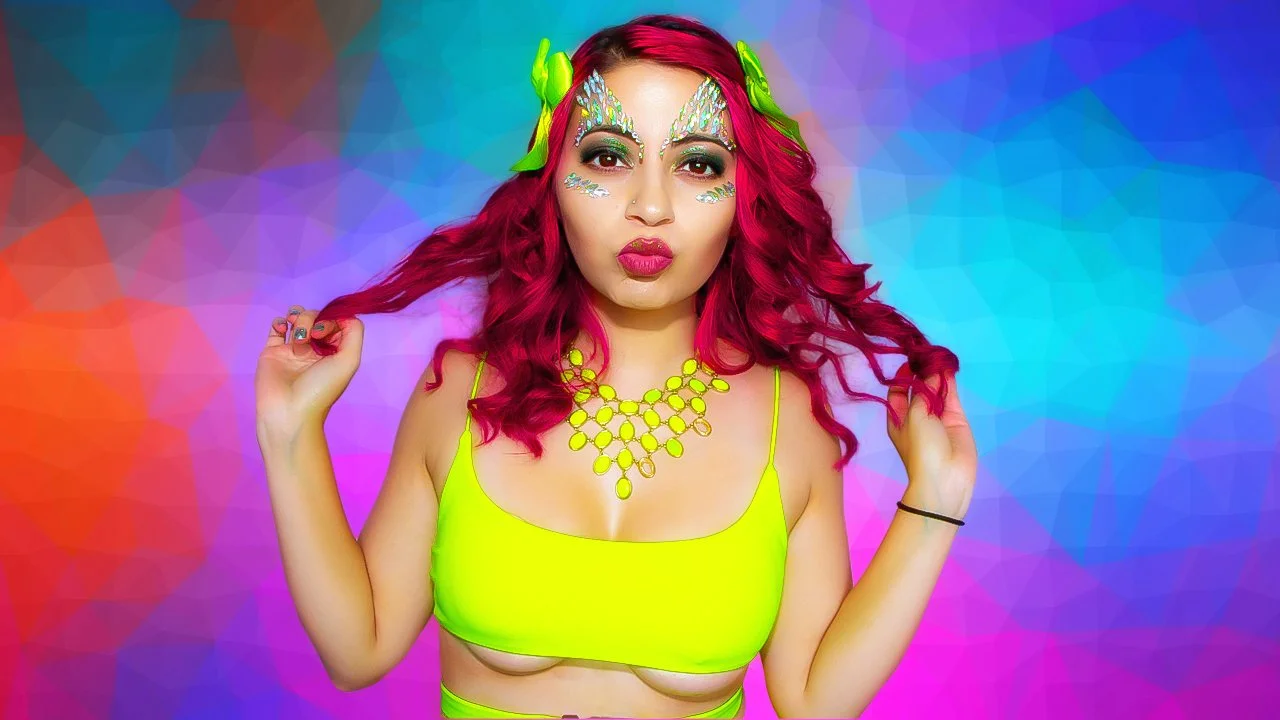 A woman with bright pink hair and butterfly makeup with rhinestones. She is wearing a neon yellow top and a matching necklace. She is holding her hair with both hands and doing a lip pout against a colorful, geometric background.