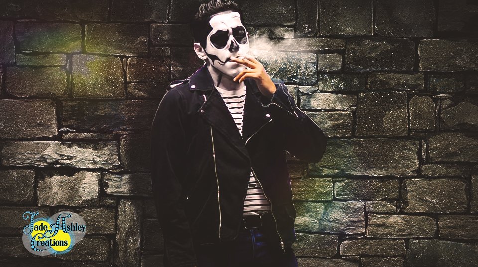 A person with a skull face paint or mask, wearing a black jacket and striped shirt, smoking a cigarette against a brick wall at night.