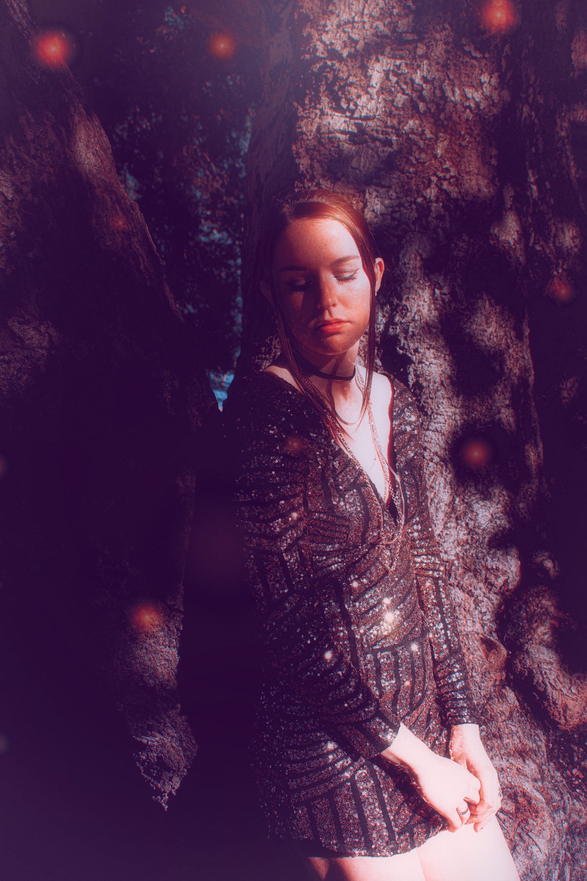 A young woman with closed eyes and red hair, wearing a black sequined dress, leaning against a tree surrounded by a dreamy, glow-like light with floating orbs.