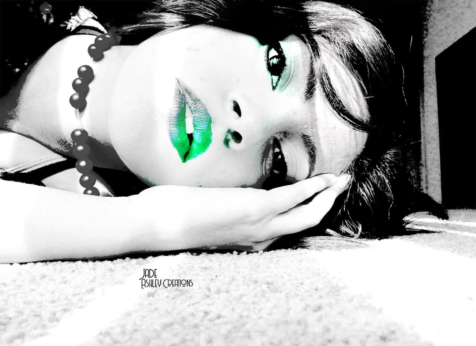 A close-up black and white photograph of a woman with long dark hair lying on the floor, with her face resting on her hand. She has digitally enhanced green lipstick and eye makeup, and is wearing a beaded necklace. There is text at the bottom left c