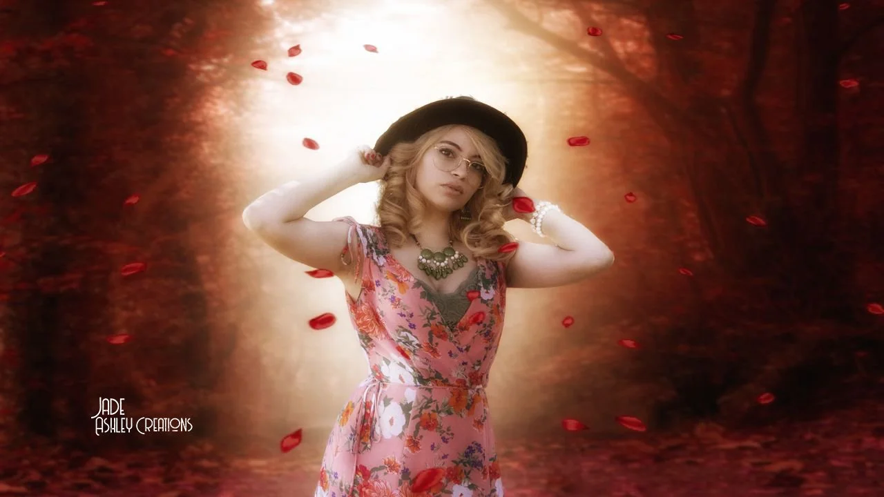 A woman with blonde curly hair wearing a floral dress, jewelry, and a black hat, standing in front of a digitally added background of a forest with red petals falling around her.
