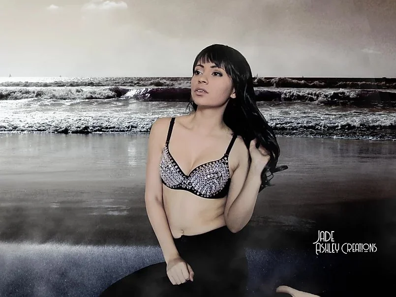 A woman with long dark hair posing in front of a beach backdrop, wearing a patterned bra and dark pants.