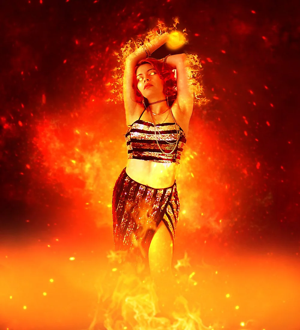 A woman with red hair in a fiery background wearing a sparkly black and silver crop top and skirt, with flames surrounding her.