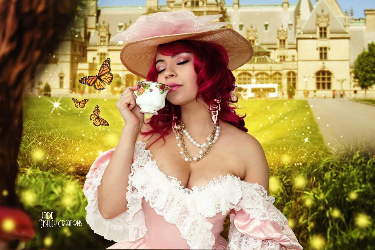 A woman with red hair wearing a pink dress, large hat, and pearl jewelry, holding a teacup with her eyes closed outdoors with a castle in the background.