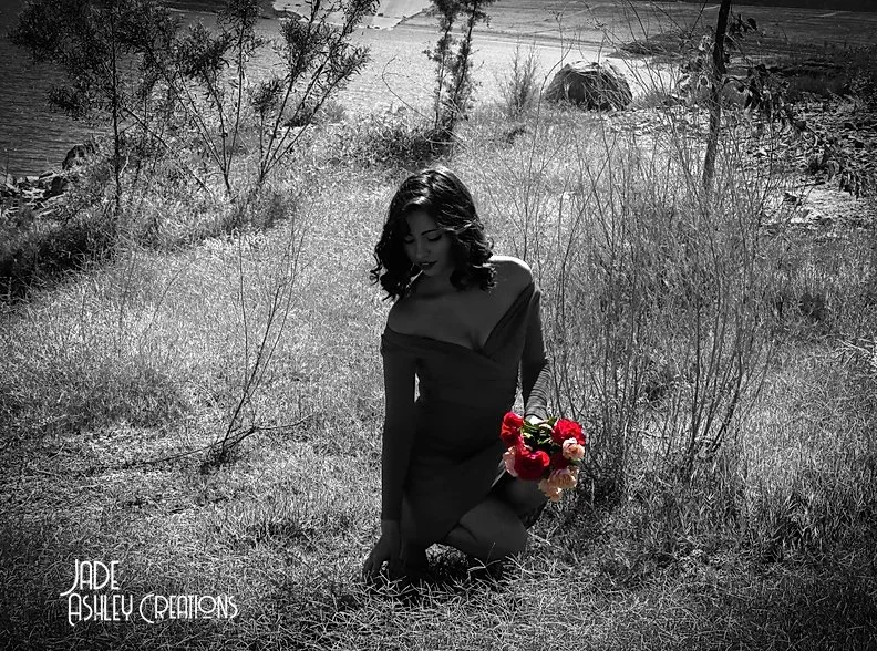 A woman with dark hair kneeling on grass near a body of water, holding a bouquet of red and pink flowers, with trees and rocks in the background, in black and white with color accents on the flowers.