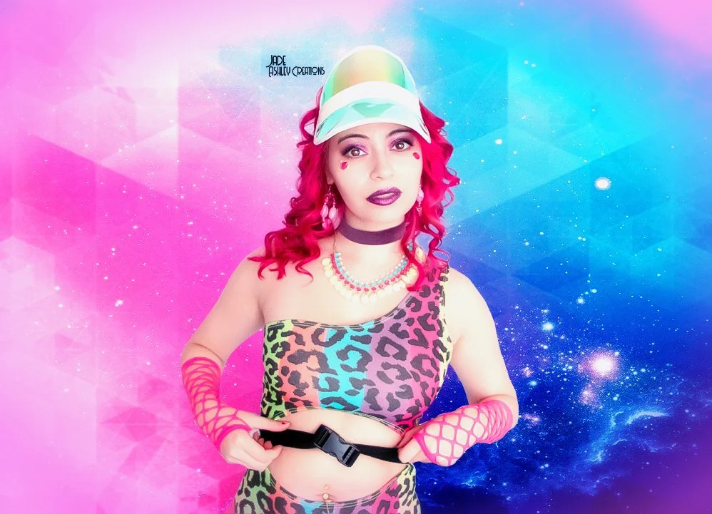 A woman with bright pink curly hair wearing a colorful bob-style cap, rainbow makeup, a leopard print crop top, and pink fishnet arm sleeves, standing in front of a vibrant, galaxy-themed background.