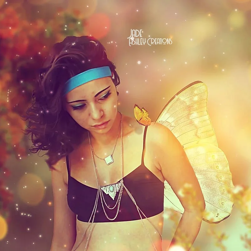 A young woman with dark, curly hair, wearing a blue headband, black crop top, and layered jewelry, has butterfly wings and a butterfly on her shoulder in a fantasy-themed photo with warm, glowing bokeh effects.