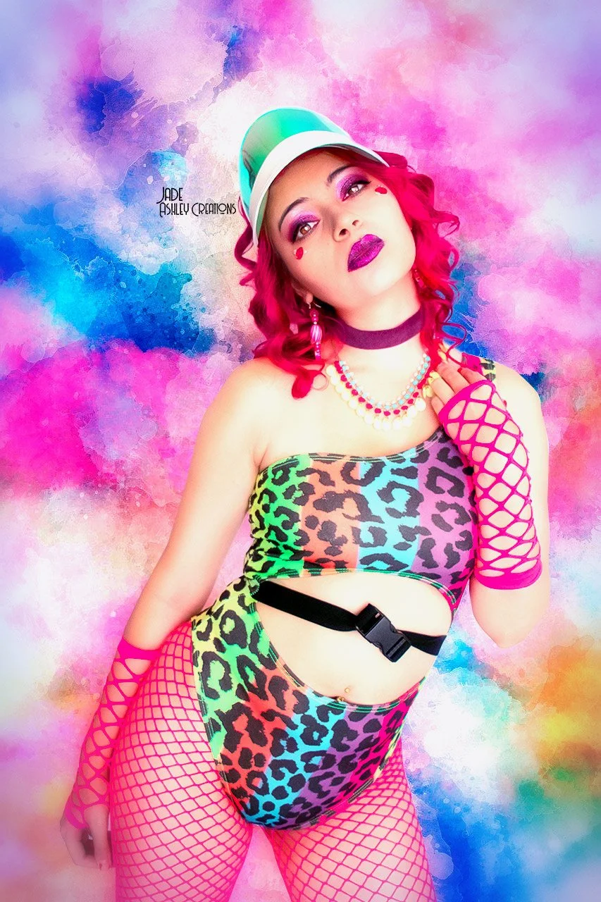 A woman with pink hair and bold makeup, wearing a rainbow-colored leopard print outfit, pink fishnet gloves, and a cap, standing against a vibrant watercolor background.
