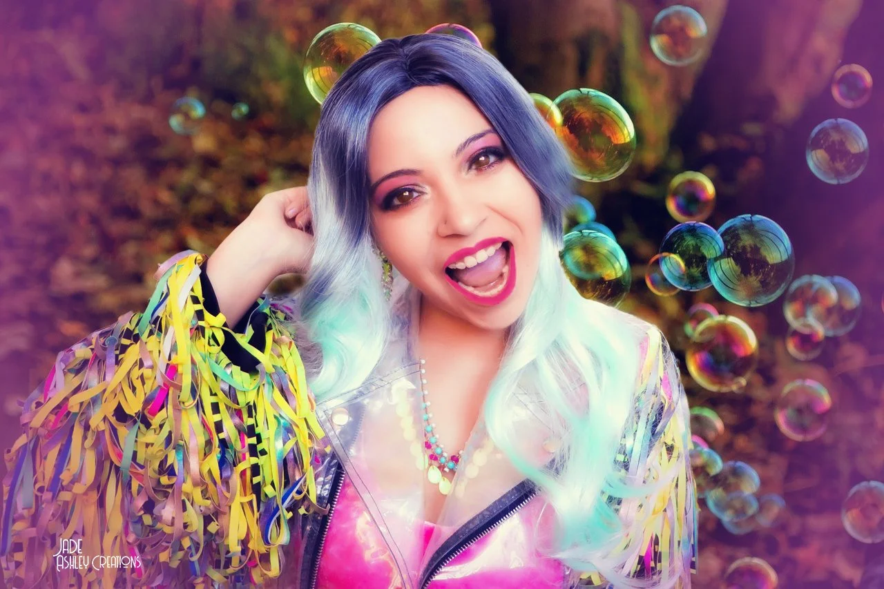 A woman with multi-colored hair, wearing a shiny, colorful jacket and a beaded necklace, is smiling and surrounded by floating bubbles outdoors.
