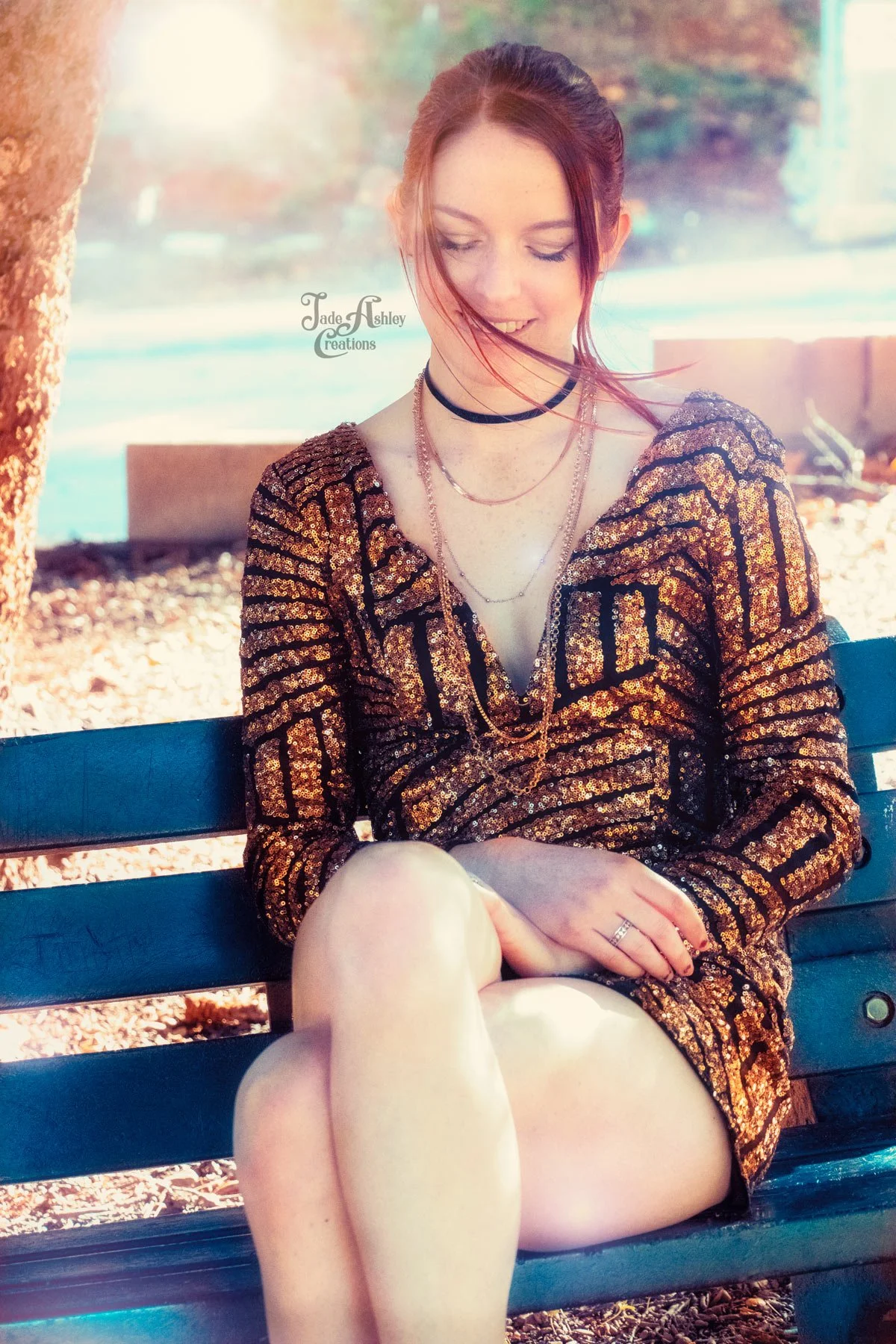 A young woman with red hair sitting on a park bench, wearing a gold and black sequin dress, black choker, layered necklaces, and rings, with her eyes looking down and smiling.
