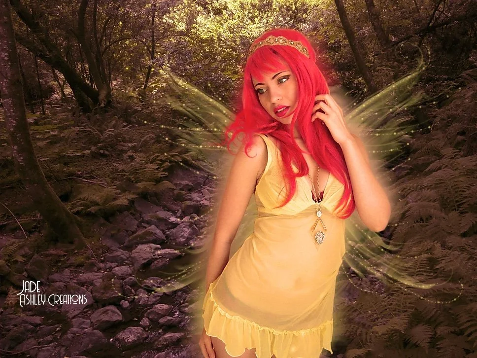 A woman with long red hair wearing a yellow dress and a gold headband, standing in a forest with glowing fairy wings and a magical aura.