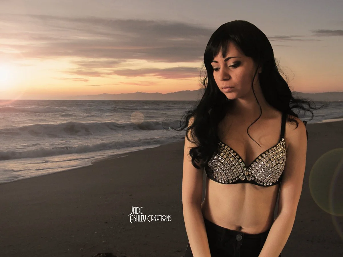 A woman with black hair and a beaded black top standing on a beach at sunset.