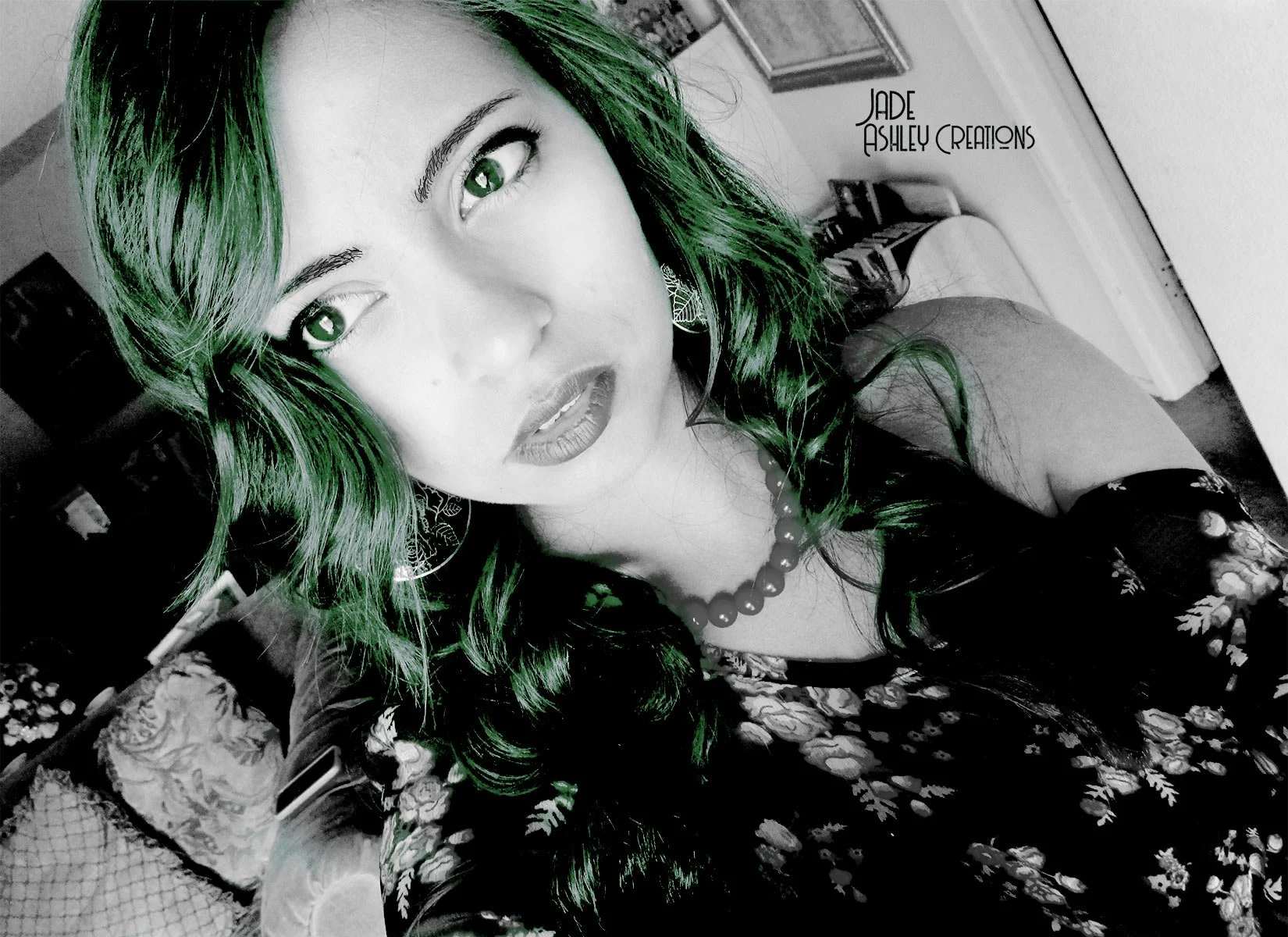 A woman with green hair and green eyes taking a selfie in a room. The photo is edited with black and white effect, except for her hair and eyes which are colored green. She is wearing a floral dress, earrings, and a beaded necklace. In the background