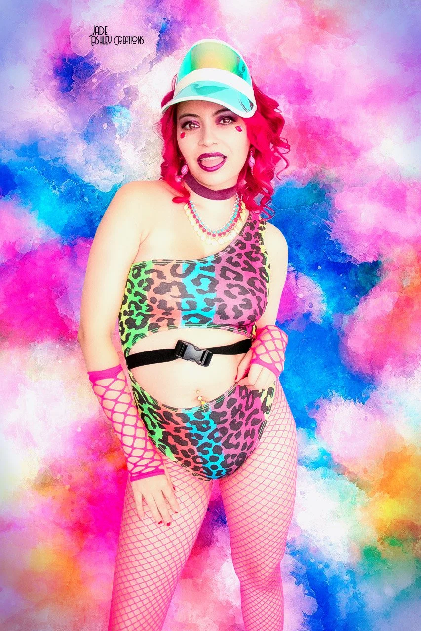Person with pink hair wearing a colorful animal print outfit, pink fishnet stockings, and a rainbow cap, standing against a vibrant, multicolored abstract background.
