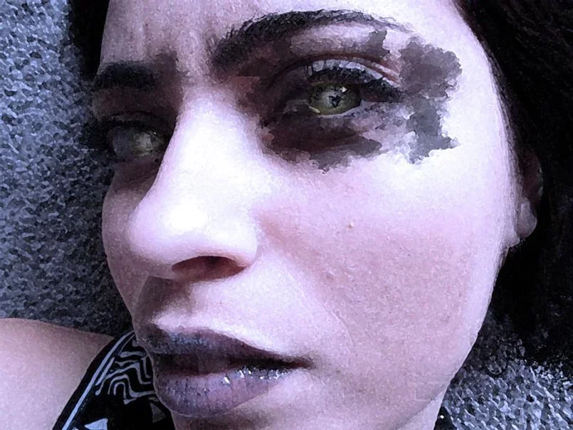 Close-up of a woman's face with dark makeup around her eyes and light-colored lips, lying on a textured surface.