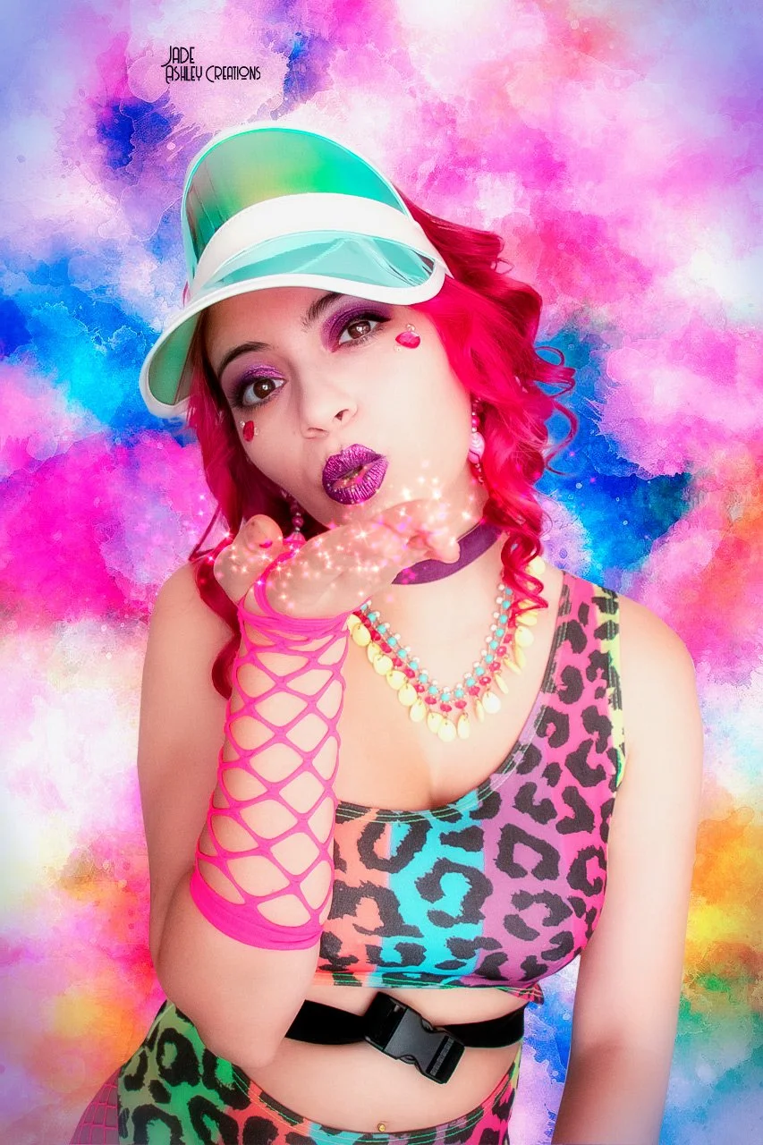 Young woman with pink hair and colorful makeup blowing glitter from her hand, wearing a vibrant animal print top, neon pink fishnet arm sleeve, layered bead necklaces, lavender lipstick, and a pink heart sticker on her cheek, against a rainbow waterc