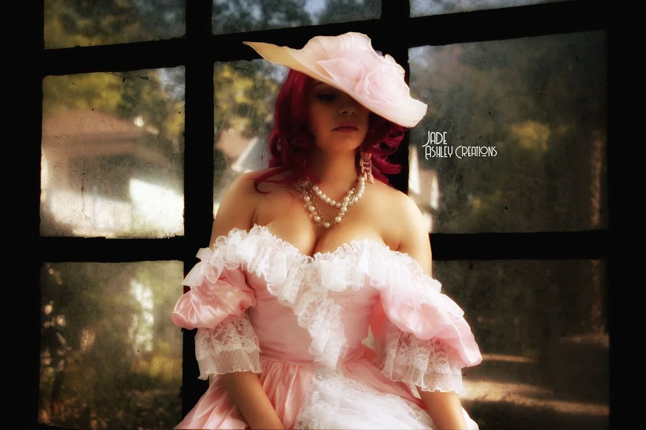 A woman with red hair wearing a pink Victorian-style dress with lace ruffles, a large pink hat, and a pearl necklace, sitting in front of a window with a blurred outdoor background.