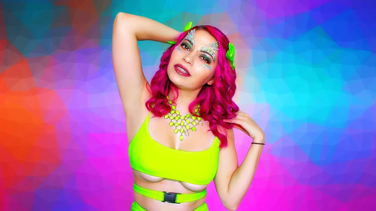 Person with bright pink hair, glittery makeup, and neon yellow outfit, posing with one arm behind head and the other touching her neck, with a colorful geometric background.