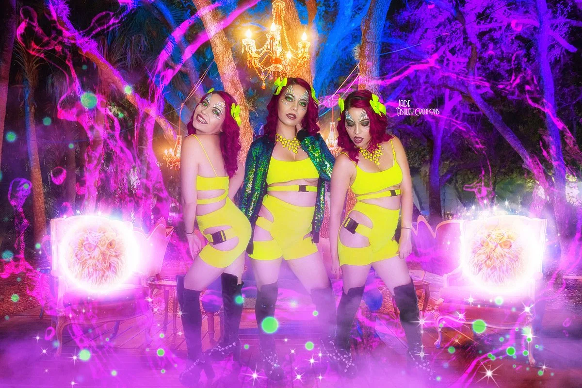 Three women in bright yellow outfits with cut-outs and thigh-high boots pose under a chandelier in a surreal, colorful setting with purple and pink neon lights, hanging trees, and glowing owl images on the chairs.
