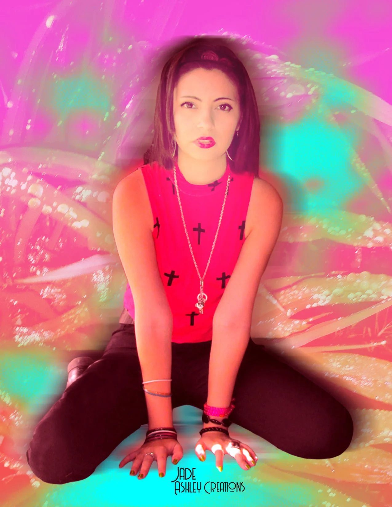 A woman with shoulder-length dark hair, wearing a pink sleeveless top with black crosses, black pants, multiple bracelets, and a long necklace with cross and key pendants, sitting on the ground against a colorful, abstract background.