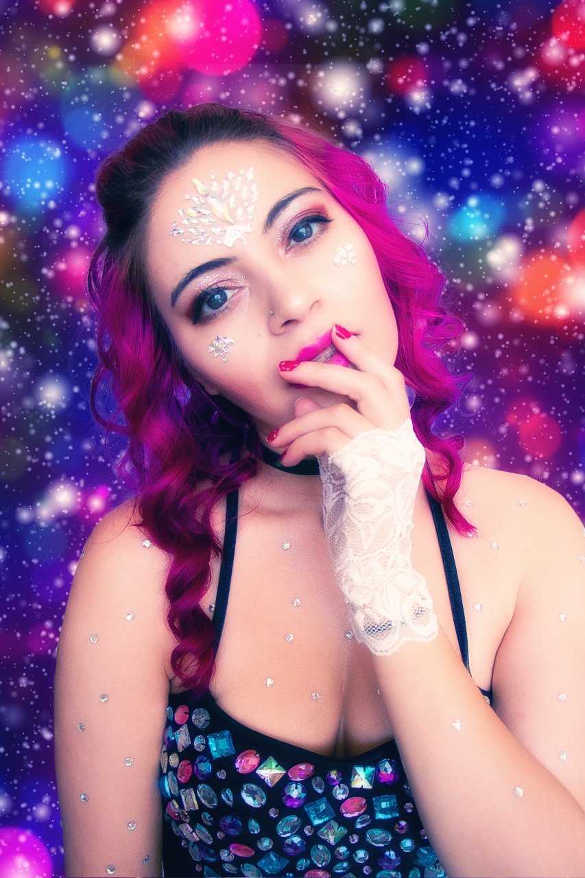 A young woman with pink and purple hair, wearing makeup, rhinestones on her face and body, and a black top covered in reflective gems, posing against a colorful, bokeh starry background.