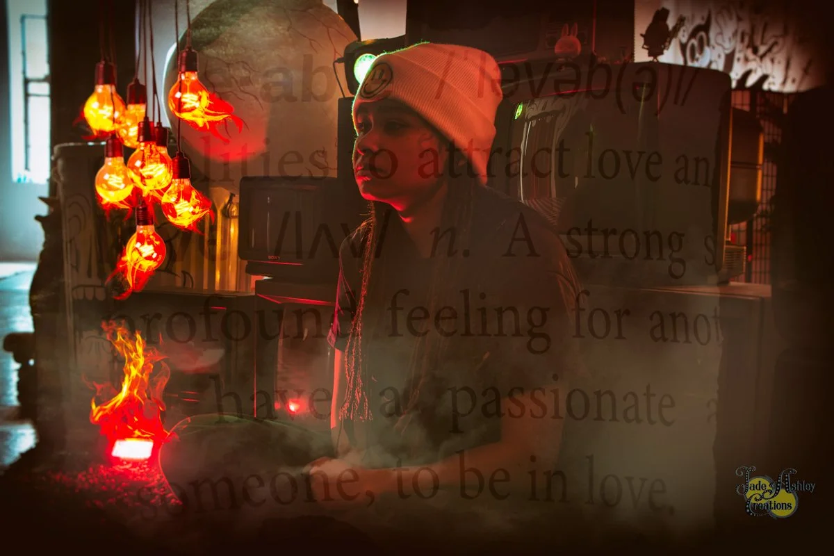 A person wearing a white knit cap sitting in a dimly lit room with red decorative lights on the left. There is text overlay on the image, and a reflection of a person working in the background.