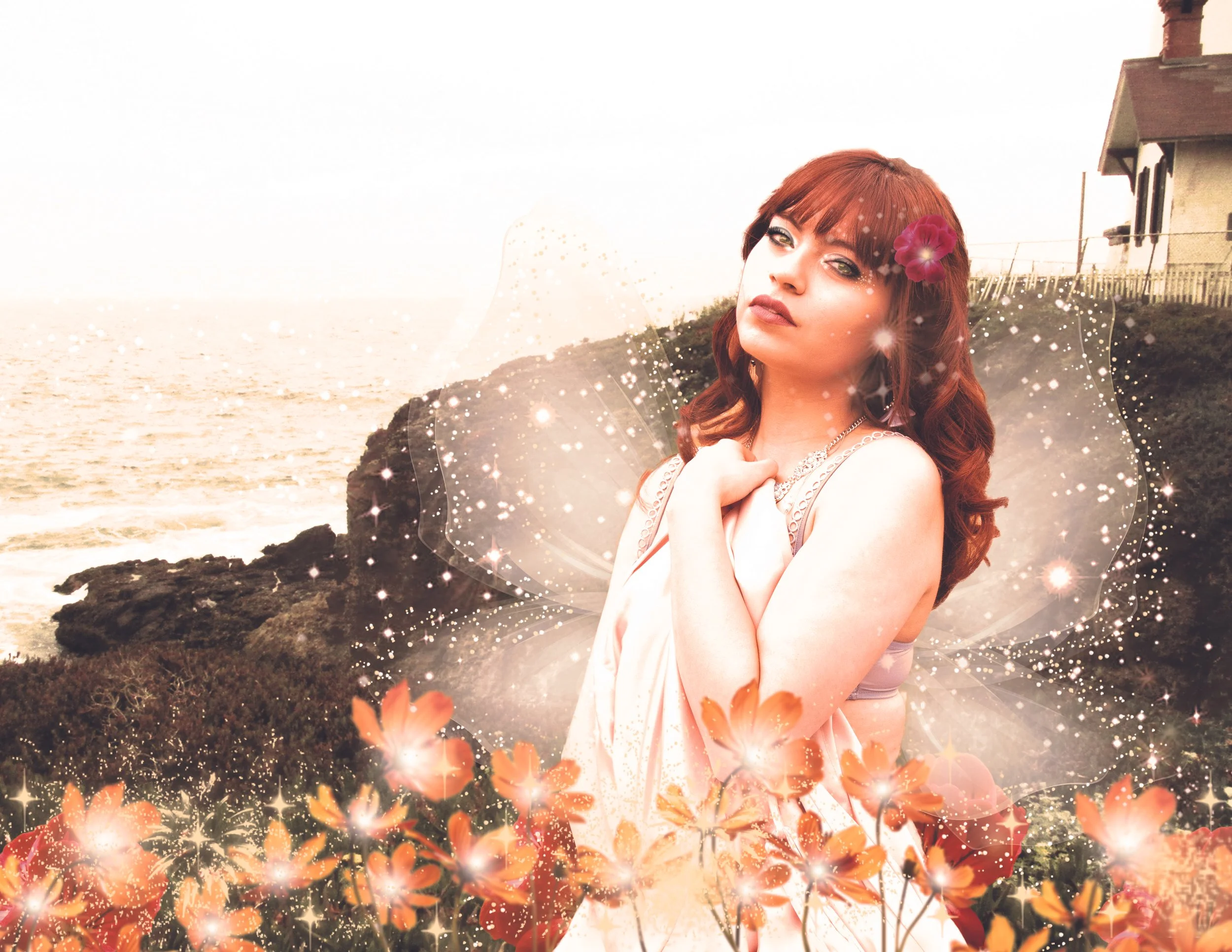 A woman with red hair and fairy wings standing on a rocky coastline near the ocean, decorated with glowing orange flowers and sparkles, with a house on a hill in the background.