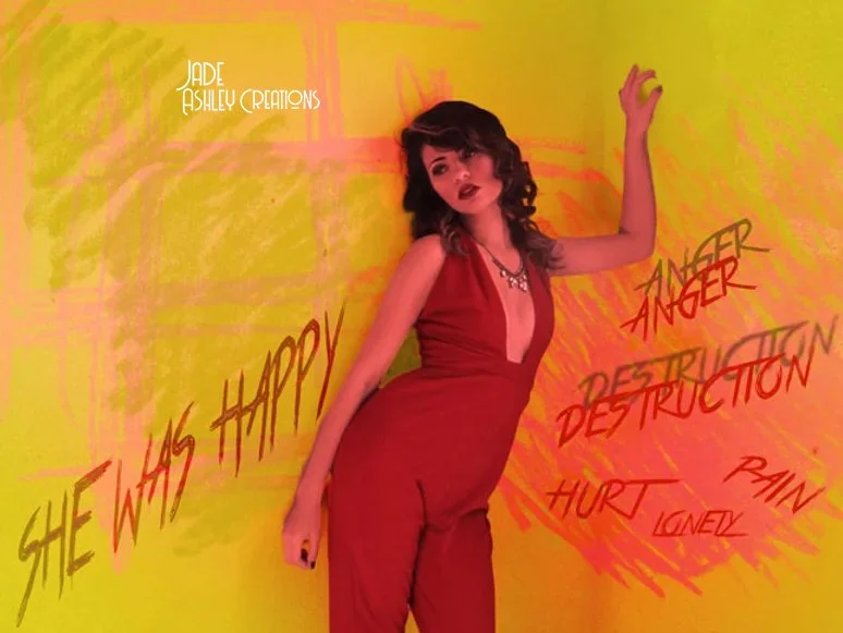 A woman with dark curly hair in a red jumpsuit standing against a yellow and orange painted background. She has her right arm raised and left arm relaxed. The image features the text 'Jade Ashley Creations' and lyrics like 'She was happy,' 'Anger,' '