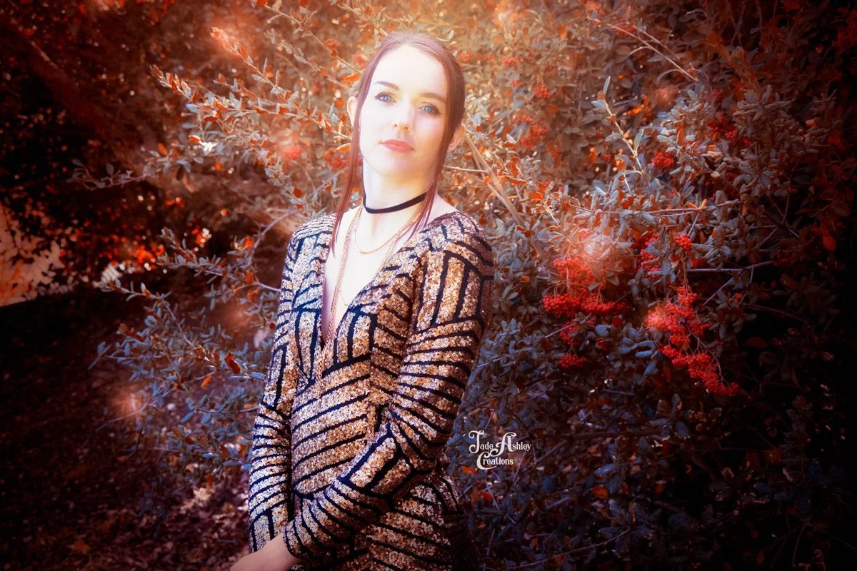 A young woman with long red hair and fair skin standing in front of a bush with red leaves and berries, wearing a black and gold patterned dress, black choker, and layered necklaces, looking at the camera with a slight smile.