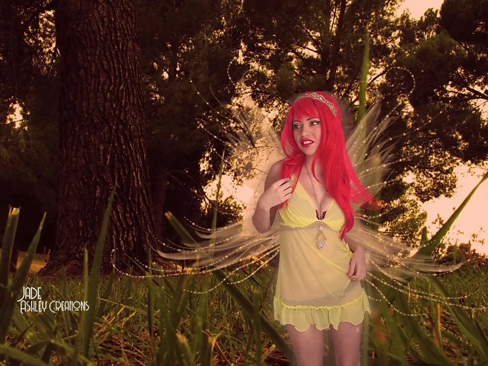 A woman with bright red hair and a tiara stands outdoors in a grassy area with trees at sunset. She is wearing a light yellow dress with ruffles and a necklace, and has fairy wings with sparkles behind her.