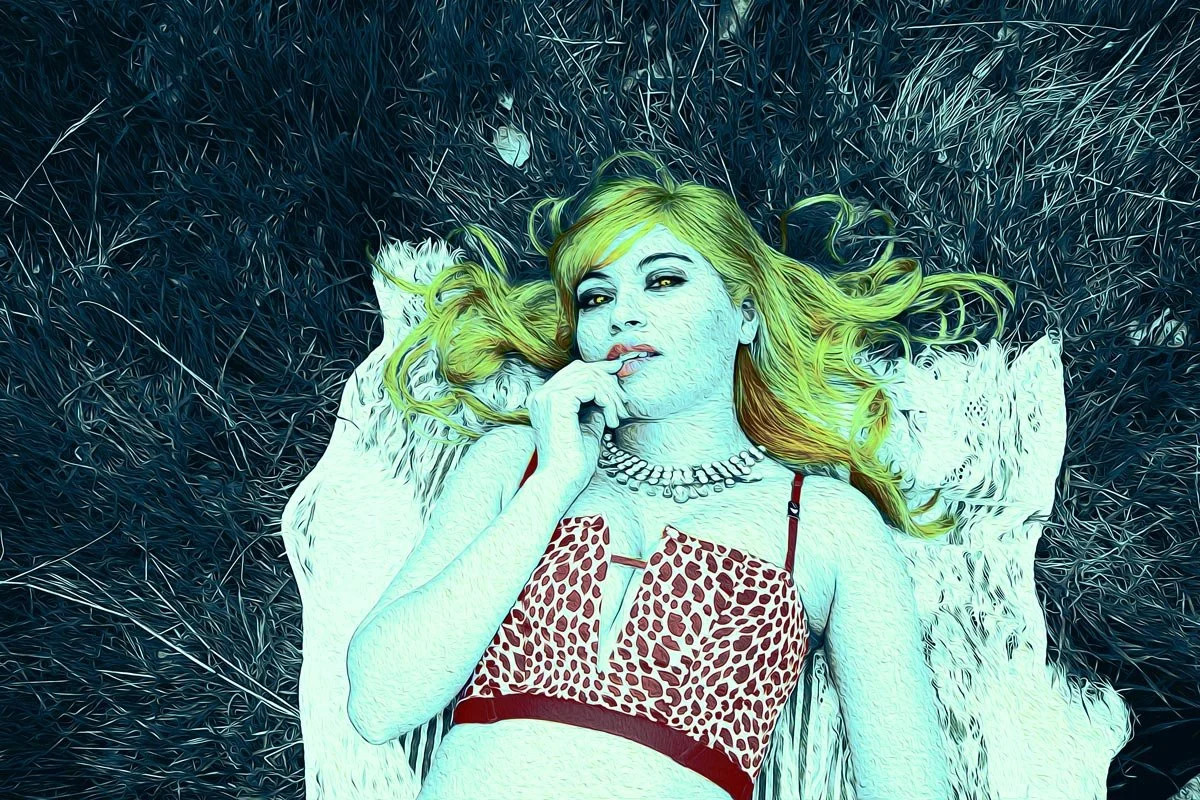 A woman with long, wavy green hair lying on a blanket on grass, wearing a leopard print top, a necklace, and touching her lips.