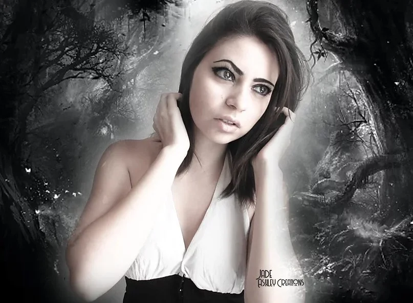 A woman with dark brown hair and makeup, wearing a sleeveless top, stands in front of a dark, moody forest background with twisted trees and fog.