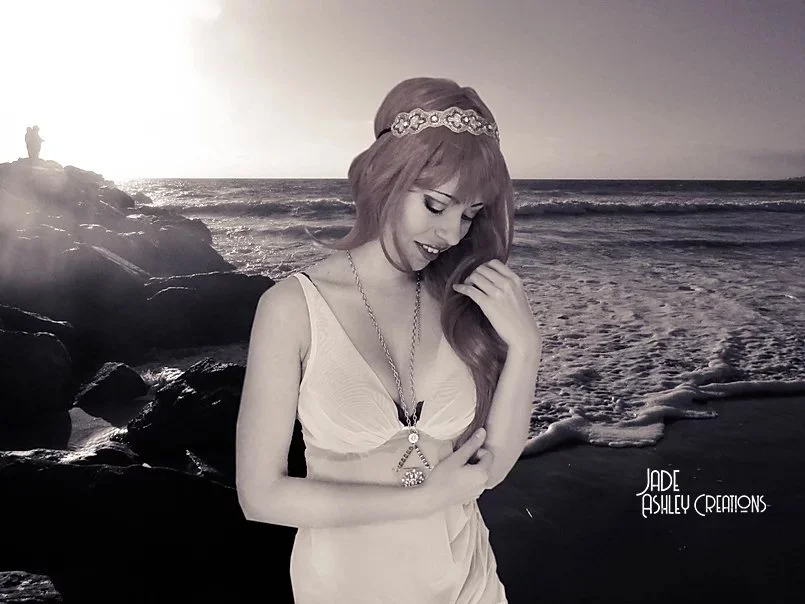 A young woman with long hair wearing a tiara and a necklace is smiling and holding her hair. She is dressed in a light-colored dress, and the background features a black-and-white beach scene with rocks and waves at sunset.