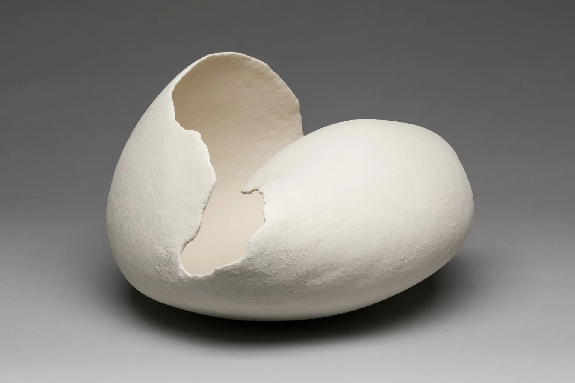 A fired porcelain paper clay sculpture of a broken white eggshell shape on a gray background.