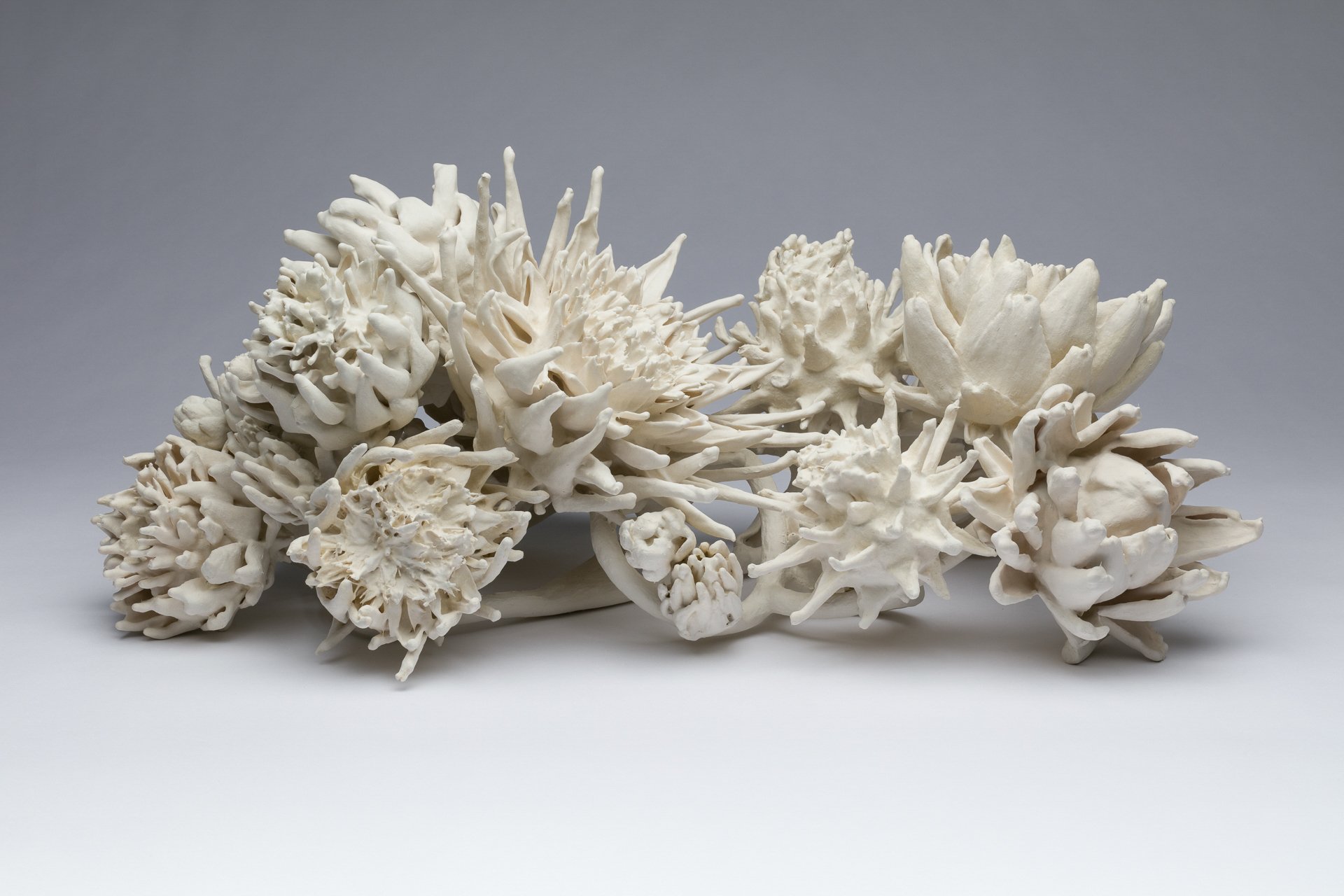 A collection of white porcelain ceramic artichokes arranged in an elongated manner on a white surface against a plain gray background.