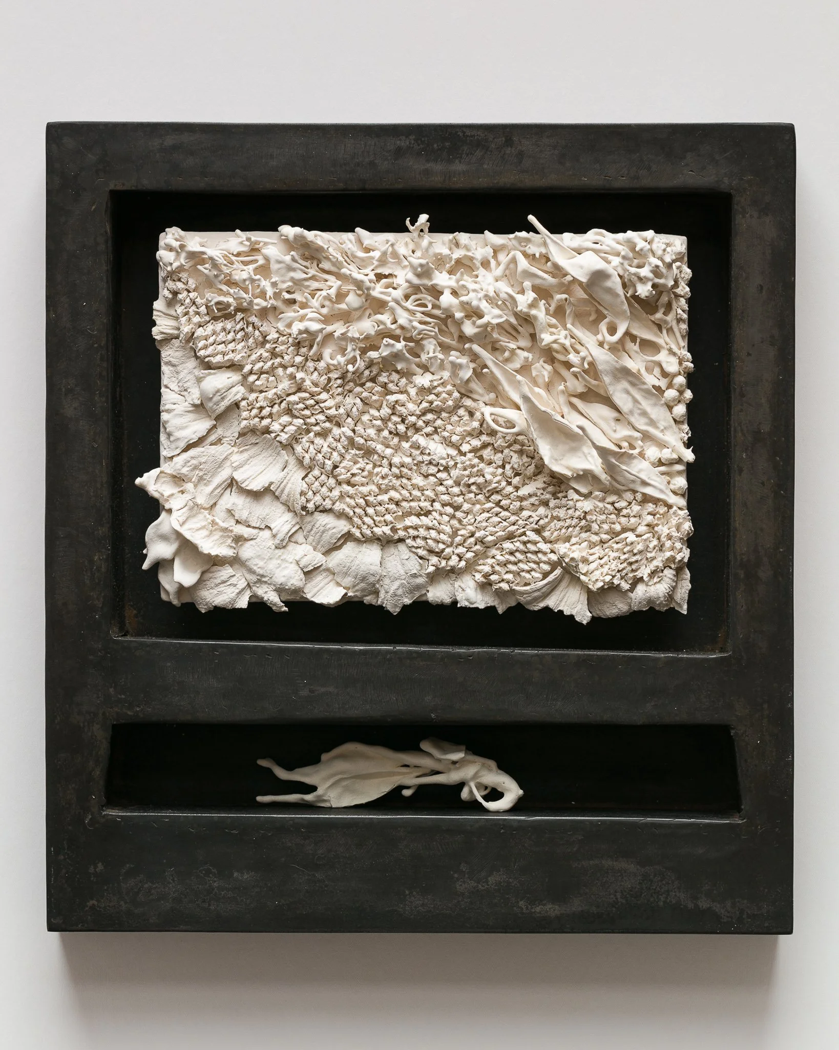 Mixed media art piece with textured white materials mounted in a black frame on a white wall.