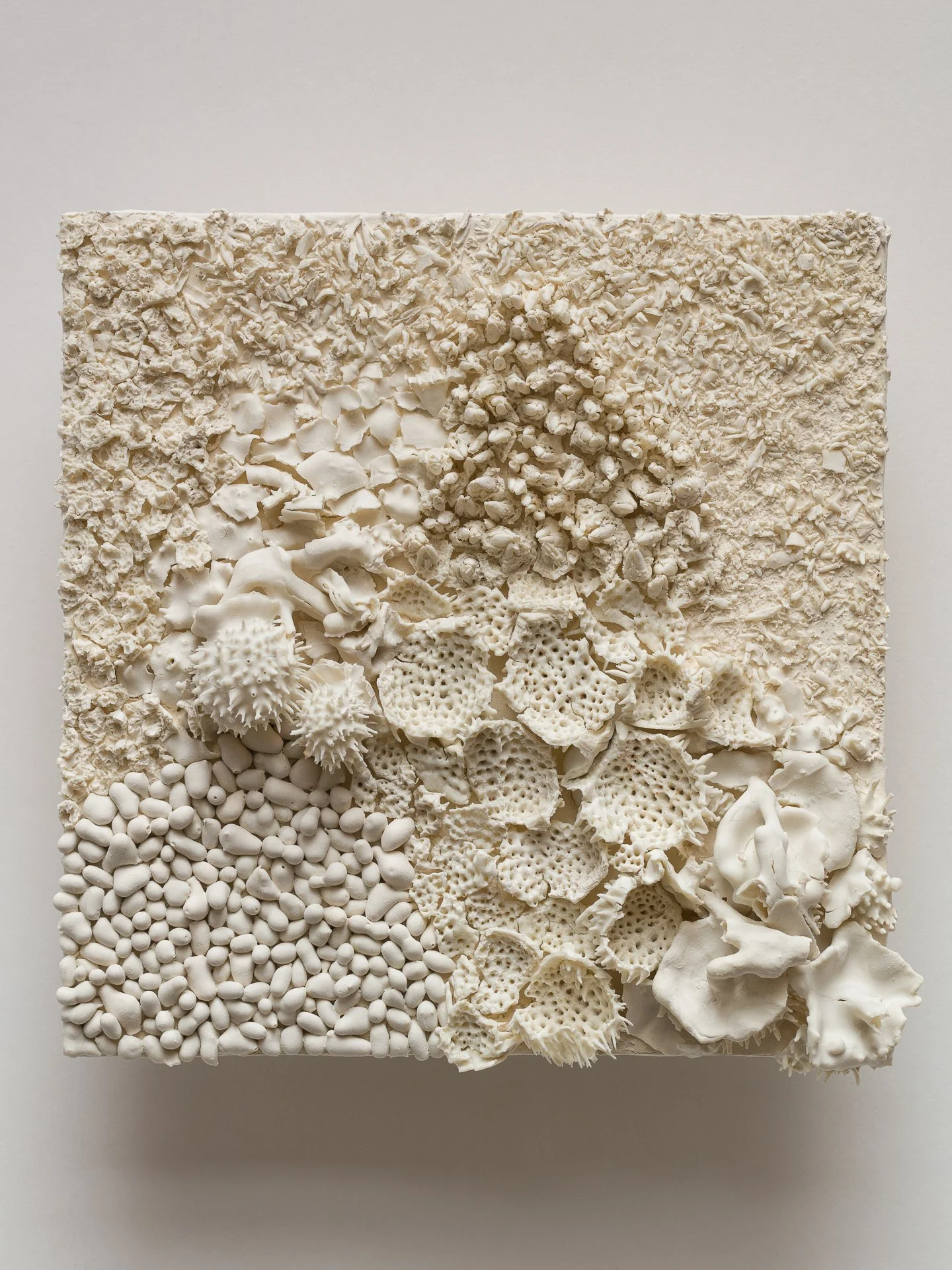 A square white art piece with various textured and patterned white materials arranged in sections.