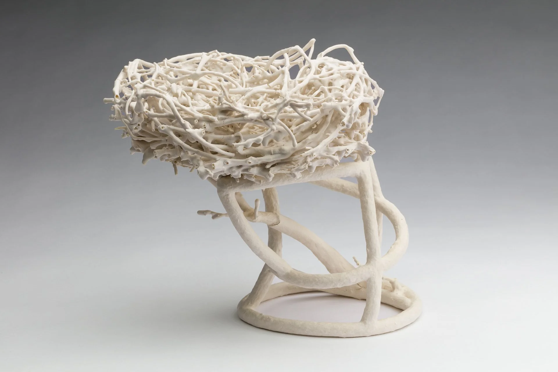 A fired porcelain paper clay sculpture resembling a nest or abstract tree, with intricate, twisted branches and a hollow, nest-like top, set against a plain grey background.