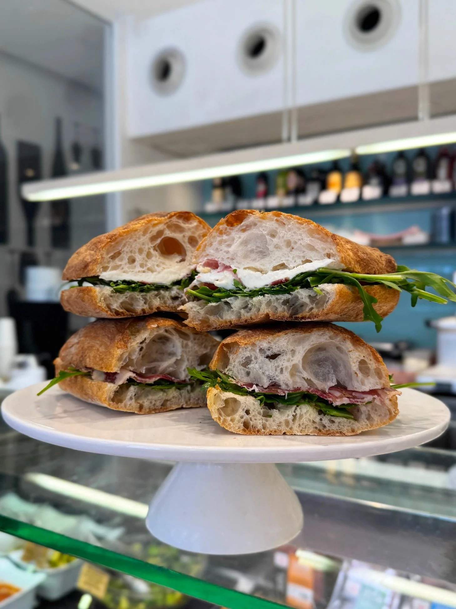 Close-up of a sandwich on a white plate, showing crusty bread, leafy greens, and slices of meat inside, with a kitchen background.