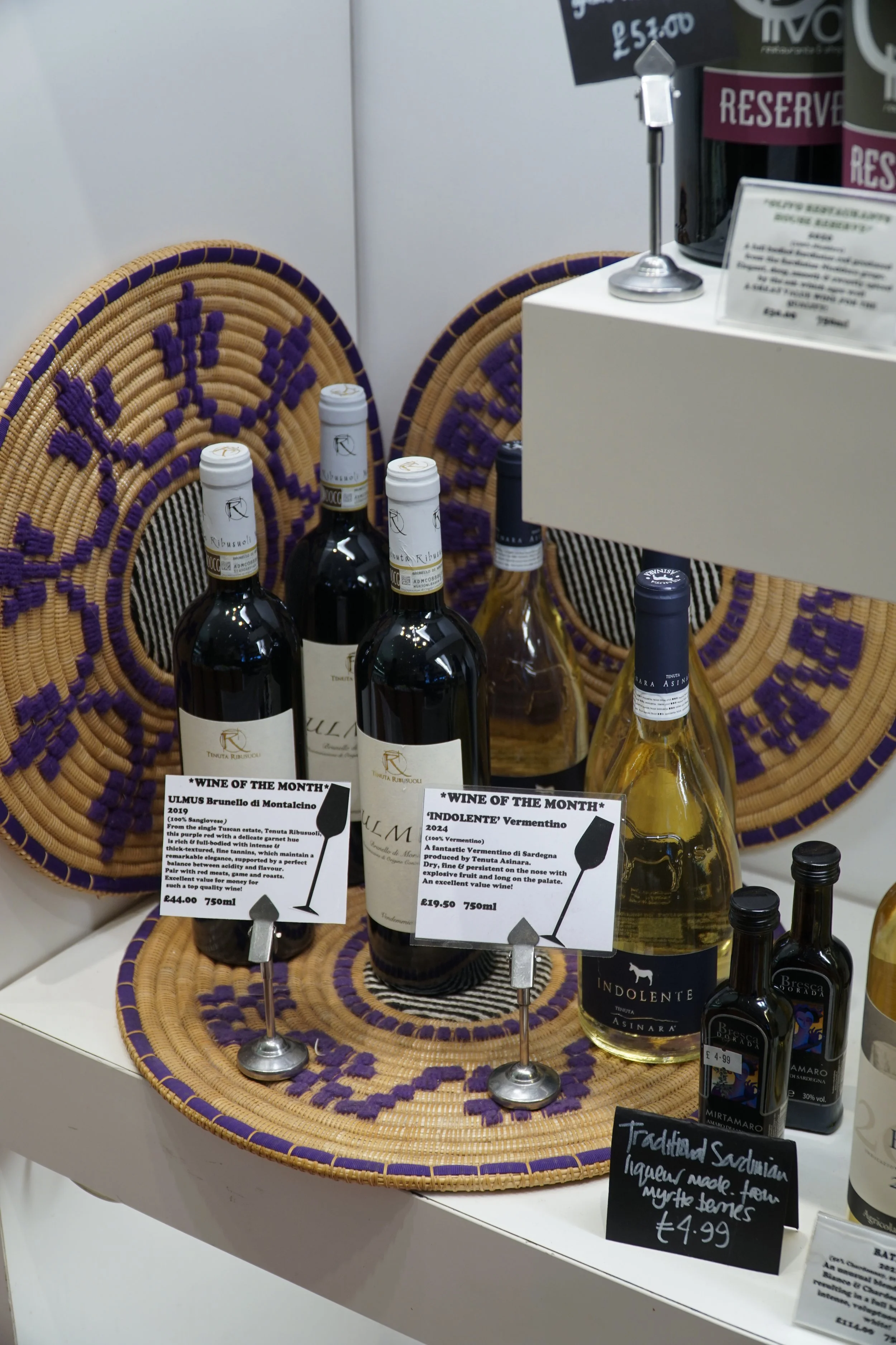 Display of wines including 'ULMUS Brunello di Montalcino' and 'INDOLENTE' Vermentino, with informational signs and woven decorative background.