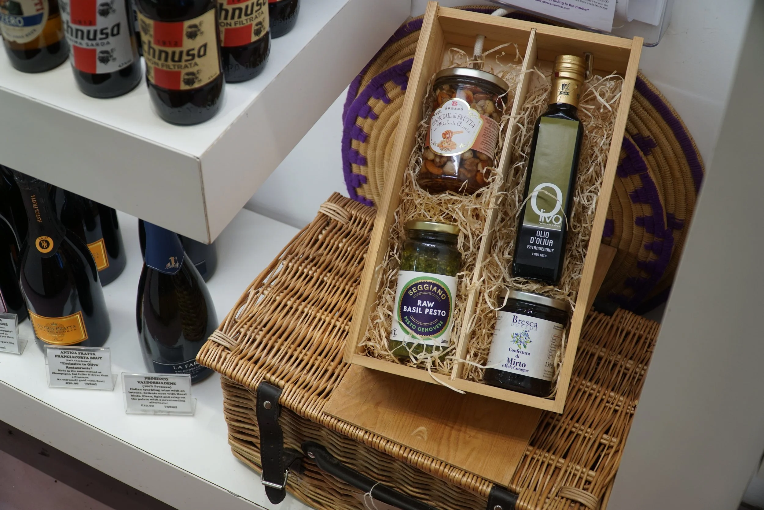 A gift box with Italian food and drink items, including jars of pesto, a bottle of olive oil, and a jar of cherry preserves, placed inside a wicker basket on a store shelf.