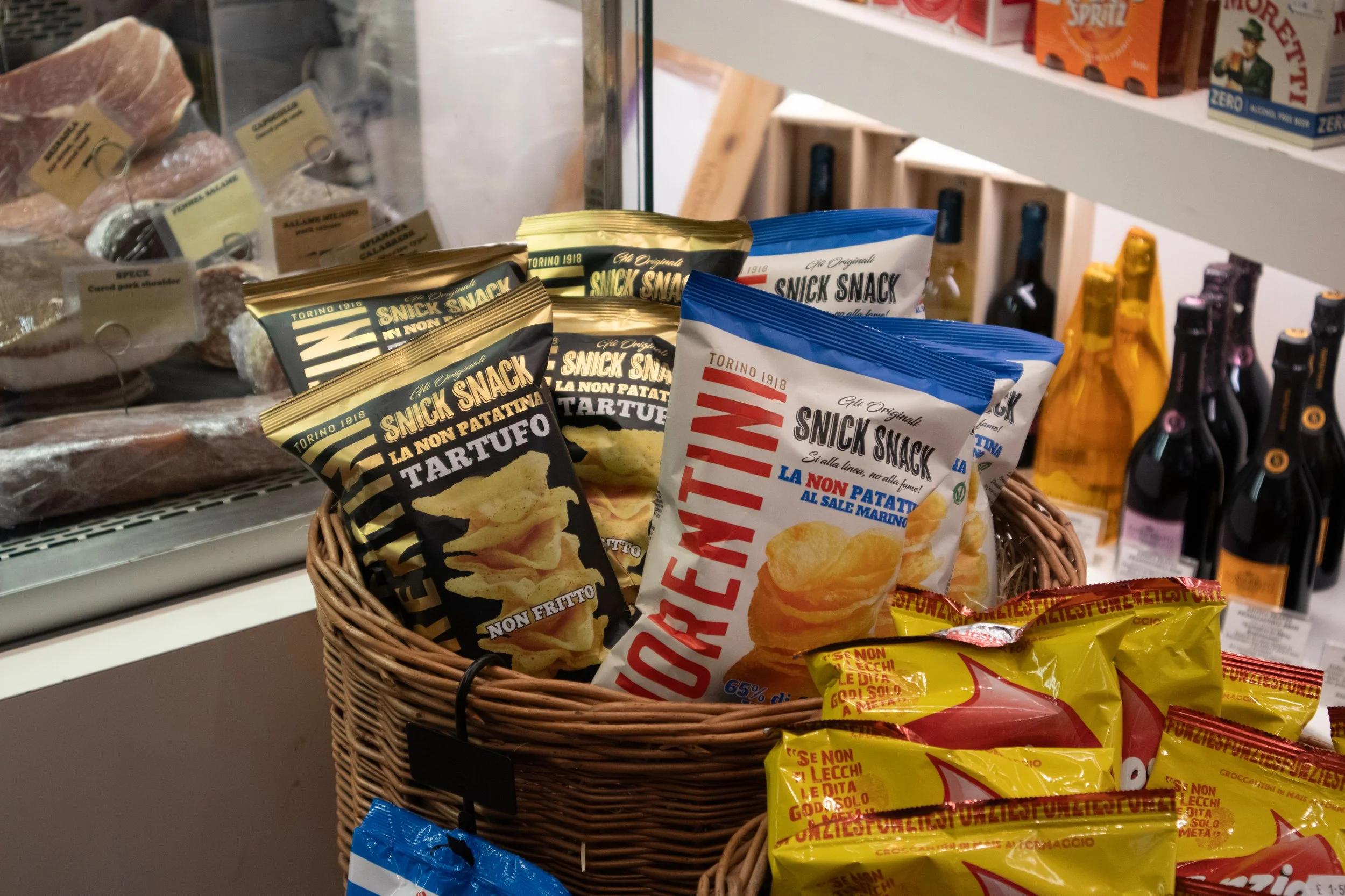 A basket filled with snack bags, including potato chips and snack sticks, in a grocery store. Behind the basket, refrigerated meat and bottles of wine are visible.