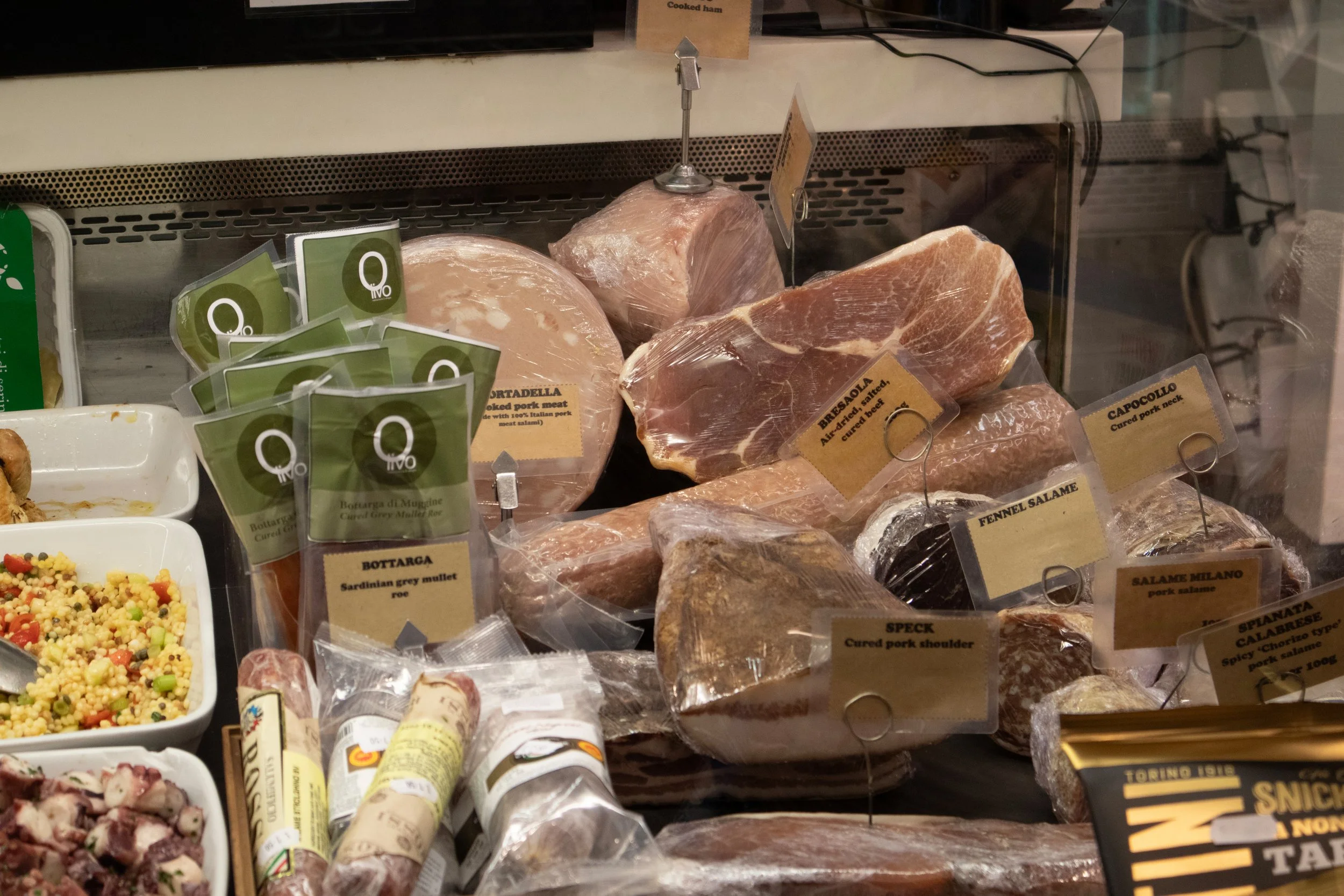 Display of various cured meats at a deli, including cured pork neck, seasoned pork, salami, and other cuts, with informational labels.