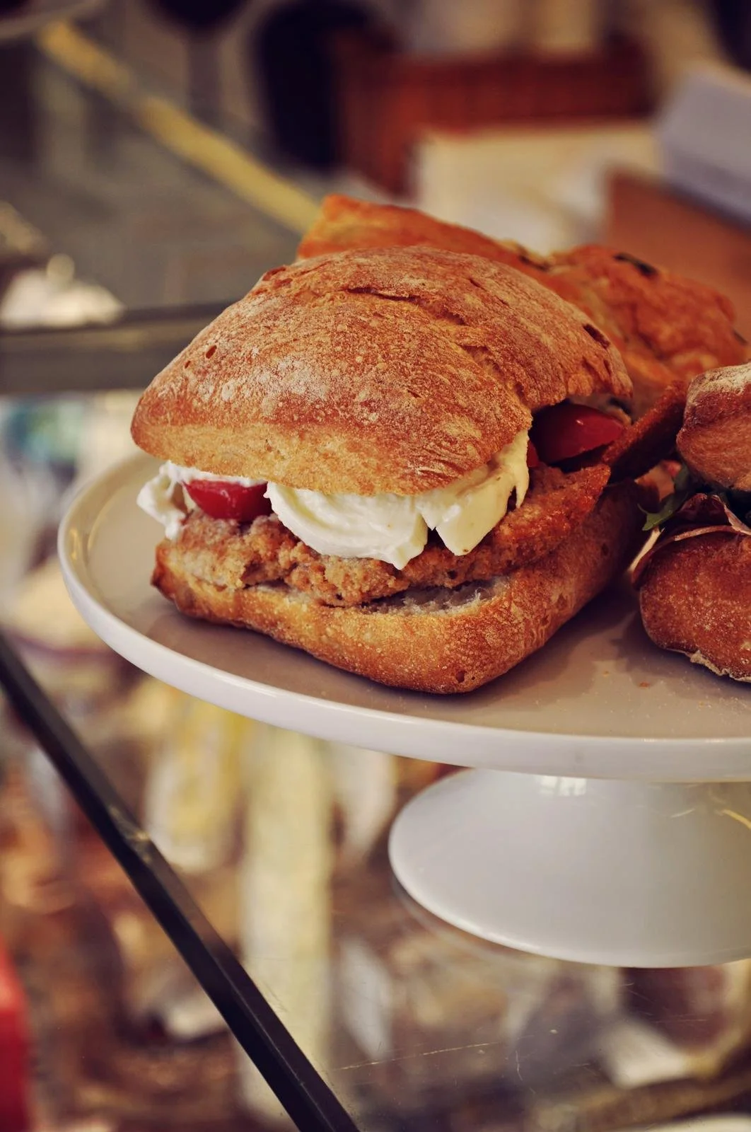 A close-up of a sandwich with a toasted bun, mayonnaise, and tomato slices, served on a white pedestal plate.