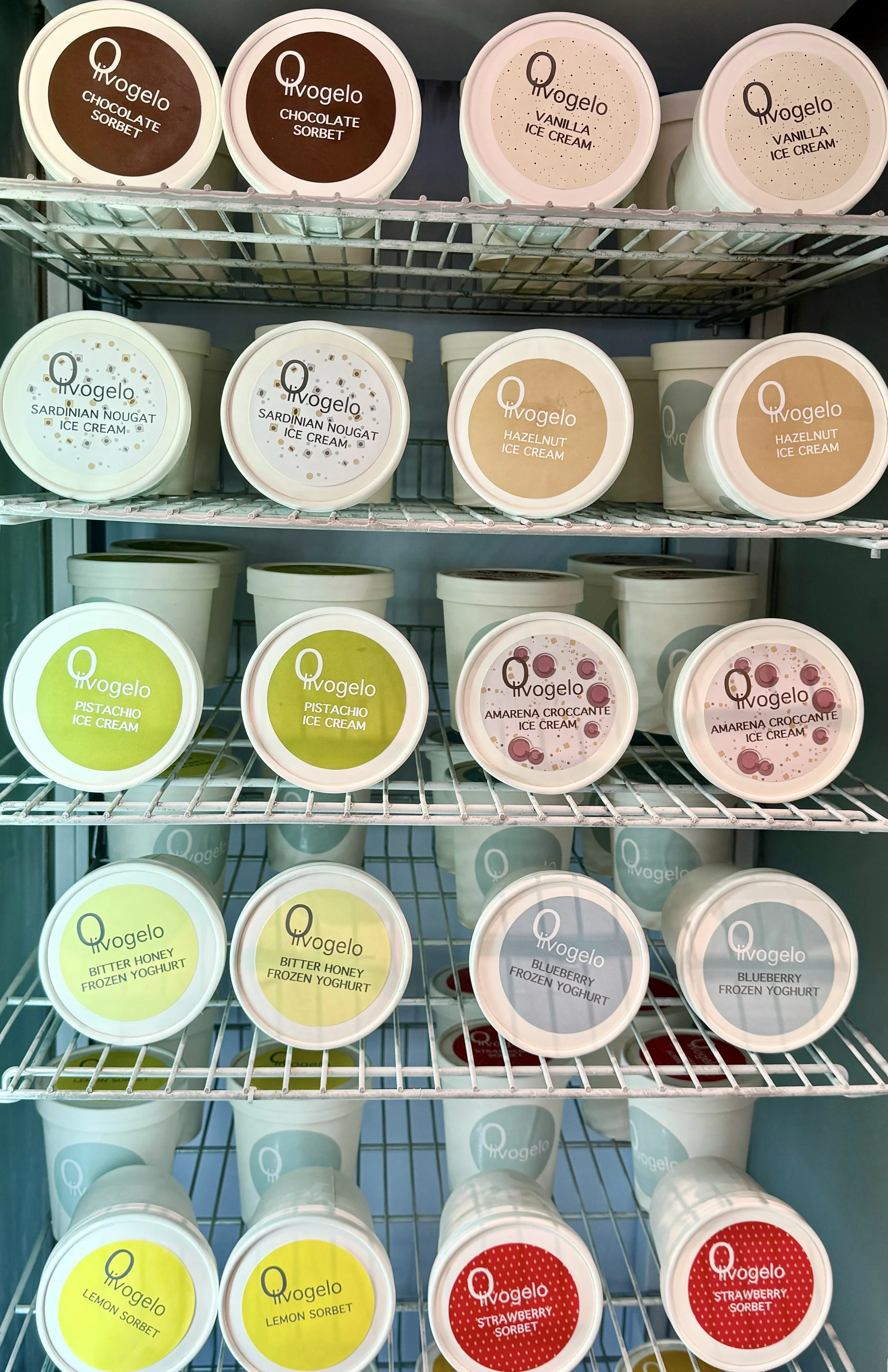 Display of various flavors of ice cream and sorbet in containers on shelves, including chocolate sorbet, vanilla ice cream, sardinian nougat ice cream, hazelnut ice cream, pistachio ice cream, amareno croccante ice cream, bitter honey frozen yogurt, blueberry frozen yogurt, lemon sorbet, strawberry sorbet, all branded with 'Ovogelo'.